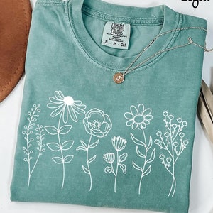 May include: A light green short-sleeve t-shirt featuring a white line art graphic of various wildflowers across the front. The shirt has a crew neckline and a Comfort Colors brand label inside the collar. The text Light green is visible.