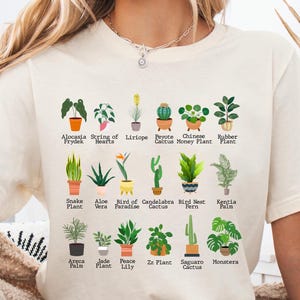 May include: A white t-shirt with a graphic of different types of houseplants, including the names of each plant. The plants are illustrated in a colorful cartoon style.