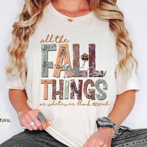 May include: A natural-colored t-shirt featuring the text "all the FALL THINGS or whatever blink 182 said" in a distressed, multi-colored block font. The design includes a skeleton graphic holding a small orange jack-o'-lantern pumpkin.