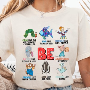 May include: Cream-colored t-shirt with colorful illustrations and the word "BE" in red. The shirt features various cartoon characters and phrases like "Calm like the Very Hungry Caterpillar" and "Kind like Rainbow Fish."