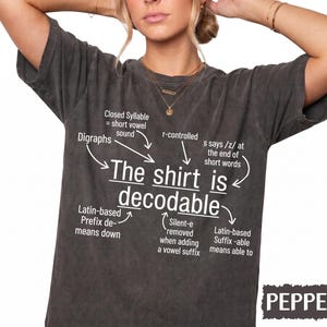 May include: A charcoal gray short-sleeve t-shirt featuring white text that reads The shirt is decodable, surrounded by linguistic annotations including Digraphs, Closed Syllable, r-controlled, s says /z/, Latin-based Prefix de-, and Suffix -able.