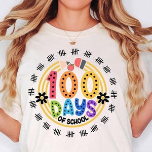 Pu&ograve; includere: Una maglietta bianca con una grafica colorata che dice "100 giorni di scuola" circondata da un cerchio giallo con segni di hash neri. La grafica include un numero 100 arcobaleno, un fiore e una matita.