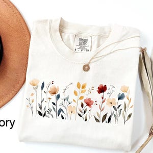 Peut inclure: T-shirt ivoire avec un motif floral &agrave; l'aquarelle sur la poitrine. Le motif pr&eacute;sente diverses fleurs dans les tons de rouge, jaune et bleu. Un collier en or avec un pendentif repose sur le t-shirt. Le t-shirt est &eacute;tiquet&eacute; "Comfort Colors".