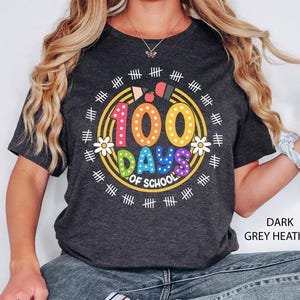 Pu&ograve; includere: Una maglietta color grigio melange scuro con una grafica colorata del testo "100 giorni di scuola" circondata da un cerchio di segni di hash e fiori.