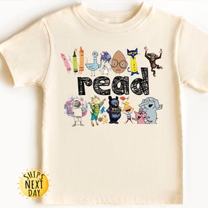 May include: Cream-colored t-shirt featuring the word "read" in black, with colorful cartoon characters and crayons. The design includes an astronaut, a bear, and a pig. A yellow sun graphic with the text "Ships Next Day" is at the bottom.