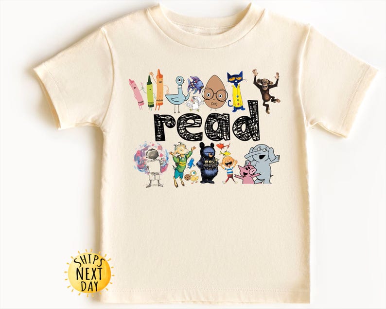 May include: Cream-colored t-shirt featuring the word "read" in black, with colorful cartoon characters and crayons. The design includes an astronaut, a bear, and a pig. A yellow sun graphic with the text "Ships Next Day" is at the bottom.
