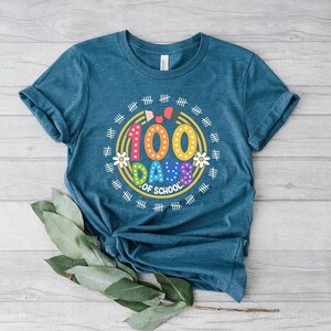 Pu&ograve; includere: Una maglietta blu turchese con una grafica colorata che dice "100 giorni di scuola" circondata da un cerchio di segni di hash e fiori.