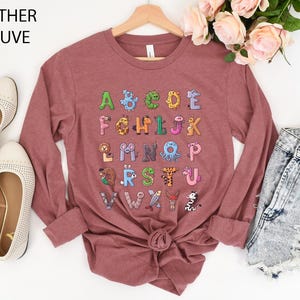 K&ouml;nnte beinhalten: Lang&auml;rmeliges Shirt in Heather-Mauve mit einem farbenfrohen Alphabet-Design mit Tierillustrationen. Das Shirt hat einen Rundhalsausschnitt und ist an der Taille gebunden. Der Text "Heather Mauve" befindet sich oben links.