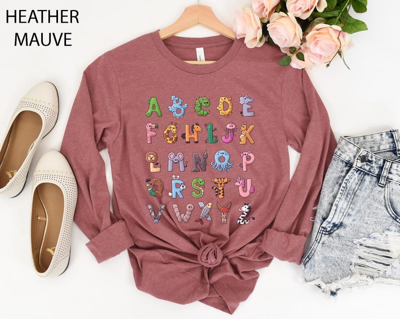 K&ouml;nnte beinhalten: Lang&auml;rmeliges Shirt in Heather-Mauve mit einem farbenfrohen Alphabet-Design mit Tierillustrationen. Das Shirt hat einen Rundhalsausschnitt und ist an der Taille gebunden. Der Text "Heather Mauve" befindet sich oben links.
