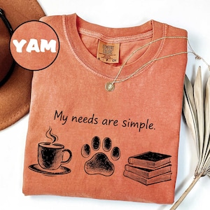 May include: A coral-colored t-shirt with the phrase "My needs are simple" printed in black. The design includes a coffee cup, paw print, and books. A gold necklace and a hat are also visible.