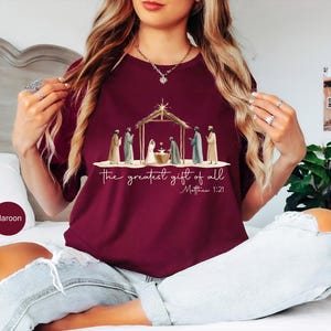 K&ouml;nnte beinhalten: Burgunderfarbenes T-Shirt mit einer Illustration der Geburt Christi, mit dem Text "the greatest gift of all" und Matth&auml;us 1:21. Die Grafik zeigt Figuren um eine Krippe unter einer Holzkonstruktion. Das Shirt ist einfarbig burgunderrot.