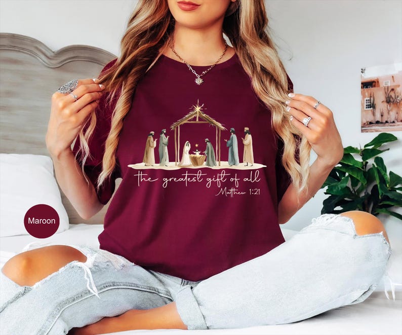 K&ouml;nnte beinhalten: Burgunderfarbenes T-Shirt mit einer Illustration der Geburt Christi, mit dem Text "the greatest gift of all" und Matth&auml;us 1:21. Die Grafik zeigt Figuren um eine Krippe unter einer Holzkonstruktion. Das Shirt ist einfarbig burgunderrot.