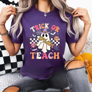 May include: A purple short-sleeve t-shirt featuring a graphic of a white ghost wearing a purple witch hat and holding a yellow pencil. The text reads TRICK OR TEACH, surrounded by small spiderwebs, bats, flowers, and a checkered pattern.