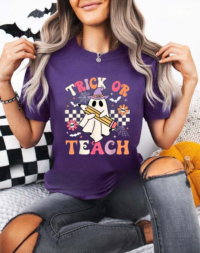 May include: A purple short-sleeve t-shirt featuring a graphic of a white ghost wearing a purple witch hat and holding a yellow pencil. The text reads TRICK OR TEACH, surrounded by small spiderwebs, bats, flowers, and a checkered pattern.