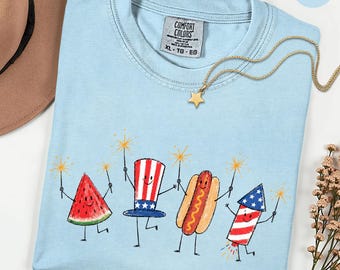 Peut inclure: Un t-shirt bleu clair avec un motif représentant une tranche de pastèque souriante, un haut-de-forme étoilé, un hot-dog et un feu d'artifice, tenant tous des cierges magiques. Le t-shirt porte une étiquette Comfort Colors et un collier étoile.