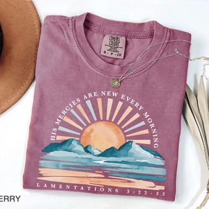 May include: Berry-colored t-shirt with the text "HIS MERCIES ARE NEW EVERY MORNING" and "LAMENTATIONS 3:22-33". The graphic features a sun, mountains, and water. A silver necklace with a pendant is on the shirt.