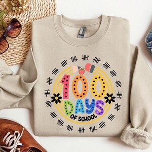 Pu&ograve; includere: Una felpa beige con una grafica colorata che dice "100 giorni di scuola" circondata da un arcobaleno e segni di hash neri.