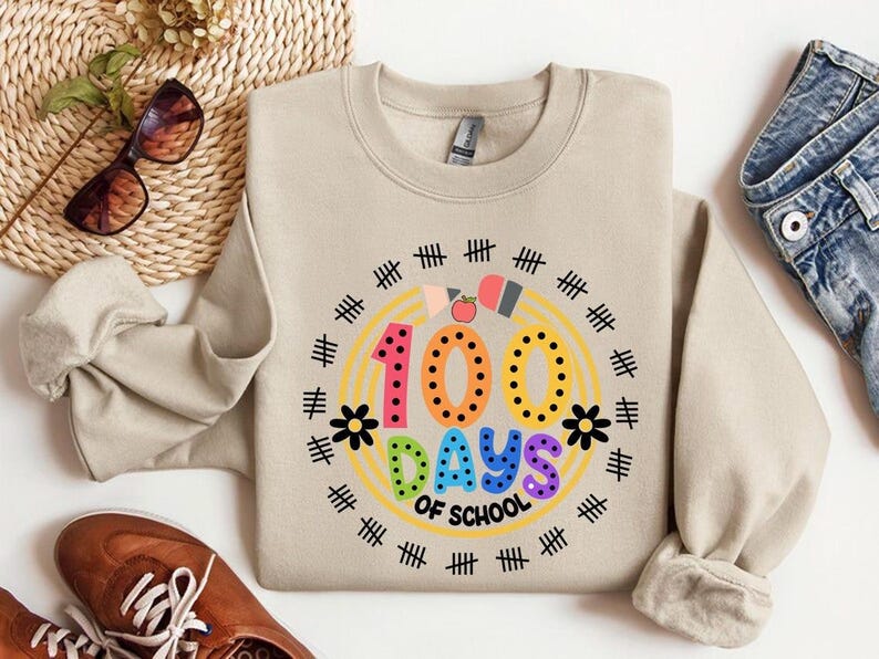 Pu&ograve; includere: Una felpa beige con una grafica colorata che dice "100 giorni di scuola" circondata da un arcobaleno e segni di hash neri.