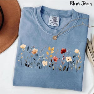 Peut inclure: Un t-shirt bleu jean avec un motif floral. Le t-shirt a un col rond et des manches courtes. Le motif floral comprend des fleurs dans les tons de rouge, jaune et blanc. Un collier en or avec un pendentif est drap&eacute; sur le t-shirt.