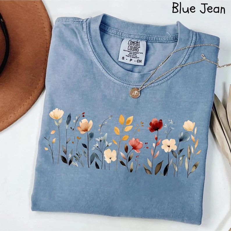 Peut inclure: Un t-shirt bleu jean avec un motif floral. Le t-shirt a un col rond et des manches courtes. Le motif floral comprend des fleurs dans les tons de rouge, jaune et blanc. Un collier en or avec un pendentif est drap&eacute; sur le t-shirt.