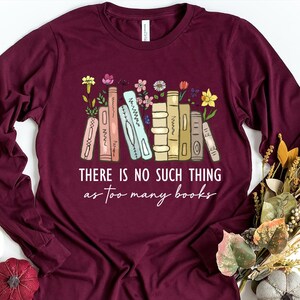 May include: Maroon long-sleeve shirt with a graphic of colorful books and flowers. The text "THERE IS NO SUCH THING as too many books" is printed below the illustration. A fall-themed floral arrangement is in the background.