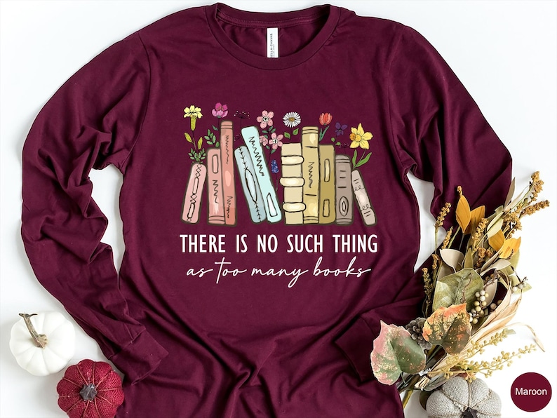 May include: Maroon long-sleeve shirt with a graphic of colorful books and flowers. The text "THERE IS NO SUCH THING as too many books" is printed below the illustration. A fall-themed floral arrangement is in the background.