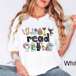 May include: White t-shirt featuring the word "READ" in black, surrounded by colorful cartoon characters and crayons. The shirt has a crew neck and short sleeves, perfect for casual wear.
