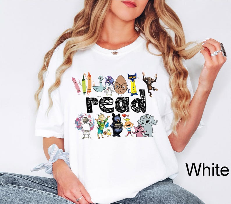 May include: White t-shirt featuring the word "READ" in black, surrounded by colorful cartoon characters and crayons. The shirt has a crew neck and short sleeves, perfect for casual wear.