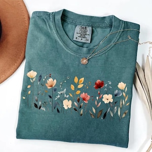 Peut inclure: T-shirt de couleur sarcelle avec un motif floral sur le devant. Le motif pr&eacute;sente des fleurs de style aquarelle dans les tons de rouge, jaune et cr&egrave;me. Un collier en or avec un pendentif repose sur le t-shirt. Le t-shirt a une &eacute;tiquette Comfort Colors.