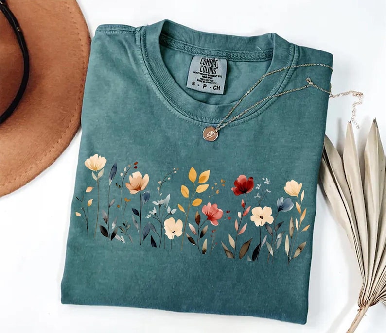 Peut inclure: T-shirt de couleur sarcelle avec un motif floral sur le devant. Le motif pr&eacute;sente des fleurs de style aquarelle dans les tons de rouge, jaune et cr&egrave;me. Un collier en or avec un pendentif repose sur le t-shirt. Le t-shirt a une &eacute;tiquette Comfort Colors.