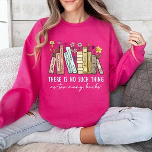 May include: A bright pink sweatshirt with the text "THERE IS NO SUCH THING as too many books." The graphic features a stack of books with flowers. The sweatshirt is long-sleeved and made of a soft material.