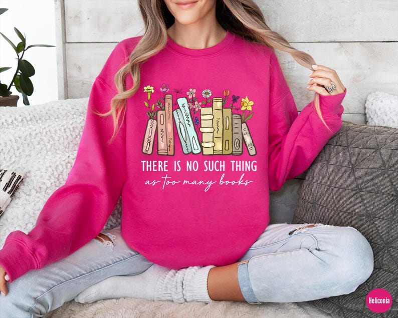 May include: A bright pink sweatshirt with the text "THERE IS NO SUCH THING as too many books." The graphic features a stack of books with flowers. The sweatshirt is long-sleeved and made of a soft material.