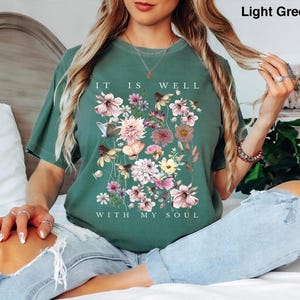 May include: A light green short-sleeve t-shirt featuring a graphic print of various colourful flowers and butterflies. The text "IT IS WELL WITH MY SOUL" is printed in white capital letters above and below the floral arrangement.