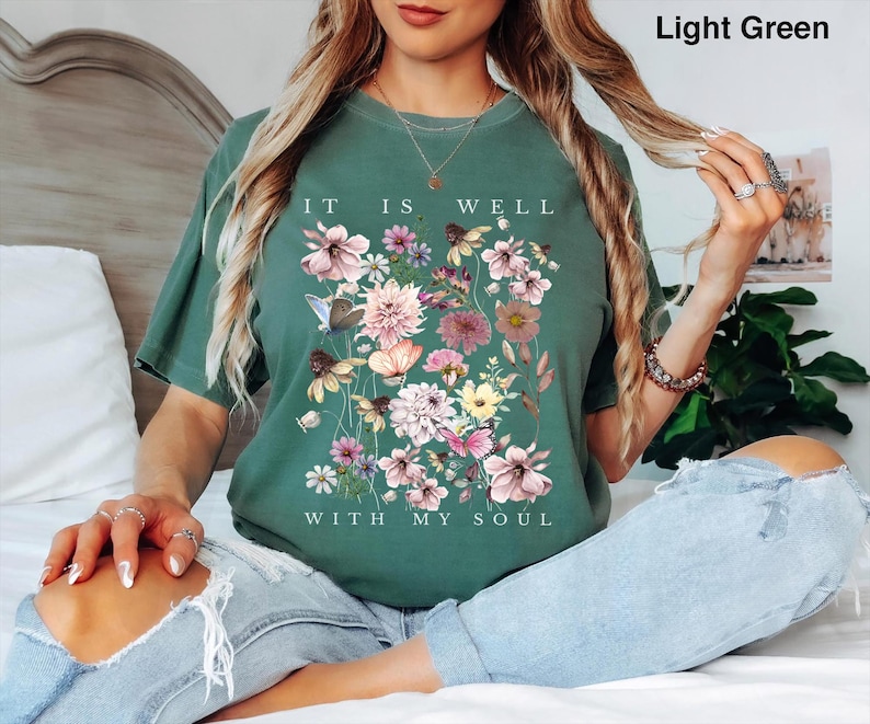 May include: A light green short-sleeve t-shirt featuring a graphic print of various colourful flowers and butterflies. The text "IT IS WELL WITH MY SOUL" is printed in white capital letters above and below the floral arrangement.