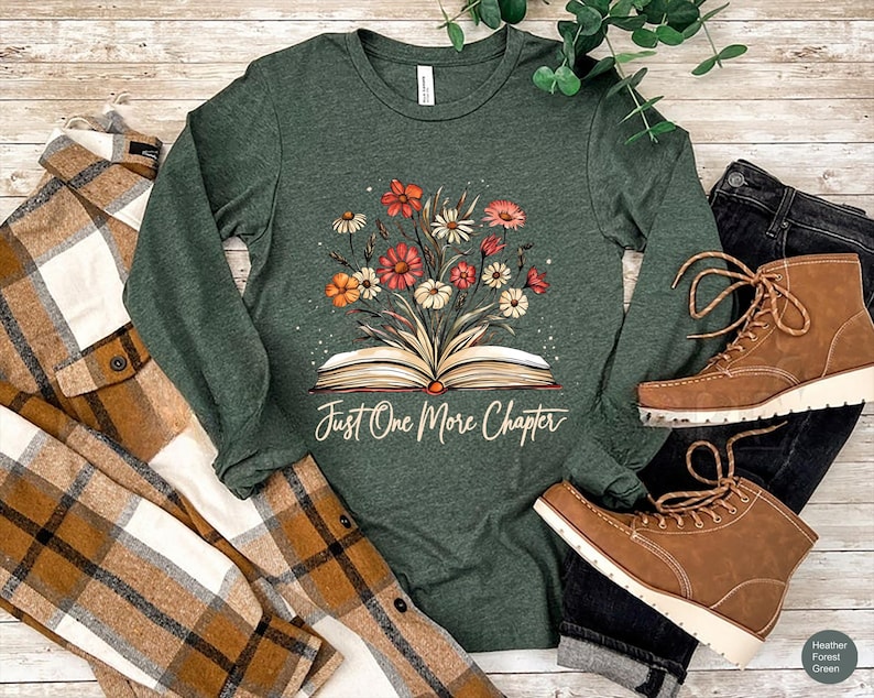 May include: A long-sleeved, heather forest green t-shirt with a floral design and an open book. The text "Just One More Chapter" is printed below the book.