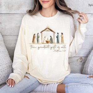K&ouml;nnte beinhalten: Cremefarbenes Langarm-Sweatshirt mit einer Aquarellillustration einer Krippenszene. Der Text "the greatest gift of all" und "Matthew 1:21" sind unter dem Bild aufgedruckt. Das Wort "NATURAL" ist oben rechts aufgedruckt.