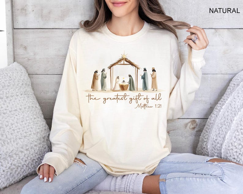 K&ouml;nnte beinhalten: Cremefarbenes Langarm-Sweatshirt mit einer Aquarellillustration einer Krippenszene. Der Text "the greatest gift of all" und "Matthew 1:21" sind unter dem Bild aufgedruckt. Das Wort "NATURAL" ist oben rechts aufgedruckt.