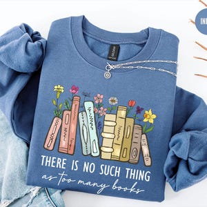 May include: Indigo blue sweatshirt with the text "THERE IS NO SUCH THING as too many books". The graphic features a stack of books with colorful flowers. A silver necklace is draped on the sweatshirt.