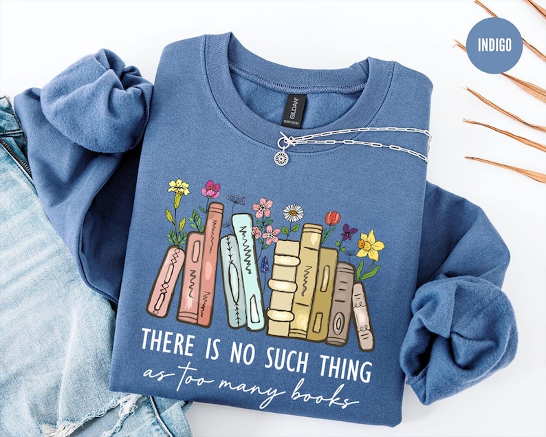 May include: Indigo blue sweatshirt with the text "THERE IS NO SUCH THING as too many books". The graphic features a stack of books with colorful flowers. A silver necklace is draped on the sweatshirt.