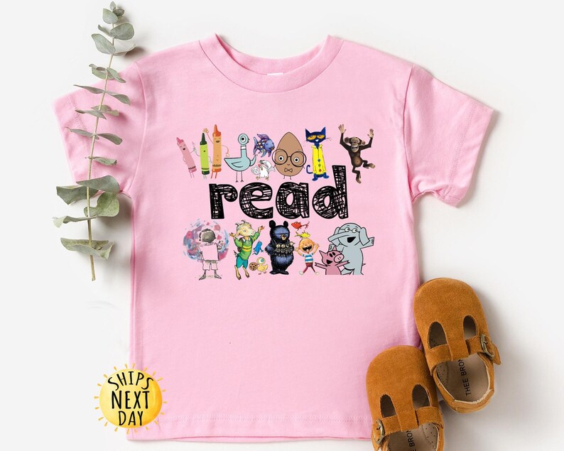May include: A pink t-shirt featuring the word "read" in black, surrounded by colorful cartoon characters and crayons. The shirt has a crew neck and short sleeves. Brown leather baby shoes are next to the shirt.