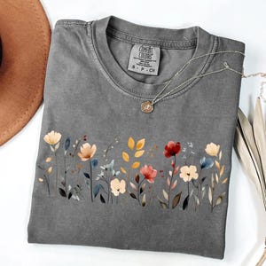 Peut inclure: Un t-shirt gris Comfort Colors avec un motif floral. Le motif pr&eacute;sente des fleurs de style aquarelle dans des tons de rouge, de jaune et de bleu. Un collier argent&eacute; avec un petit pendentif repose sur le t-shirt. Le t-shirt est pli&eacute;.