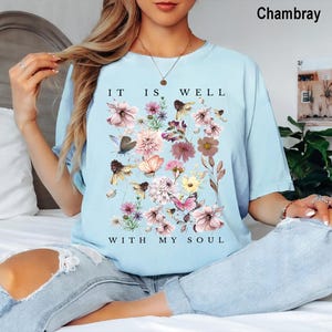 May include: A light blue short-sleeved t-shirt featuring a graphic print of various flowers, butterflies, and bees. The text "IT IS WELL WITH MY SOUL" is printed in black capital letters above and below the floral arrangement on the front.