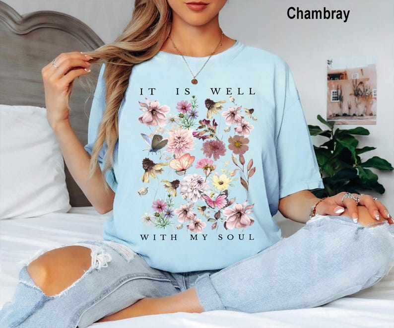 May include: A light blue short-sleeved t-shirt featuring a graphic print of various flowers, butterflies, and bees. The text "IT IS WELL WITH MY SOUL" is printed in black capital letters above and below the floral arrangement on the front.