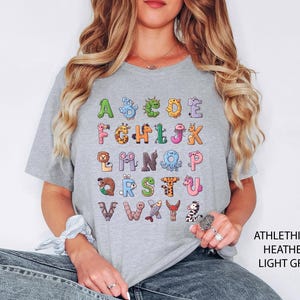 K&ouml;nnte beinhalten: Hellgraues T-Shirt mit einem farbenfrohen Alphabet-Design, wobei jeder Buchstabe durch eine Tierillustration dargestellt wird. Das Shirt besteht aus einem weichen Material und eignet sich f&uuml;r Freizeitkleidung. Der Text "ATHLETHICH HEATHER LIGHT GREY" ist rechts aufgedruckt.