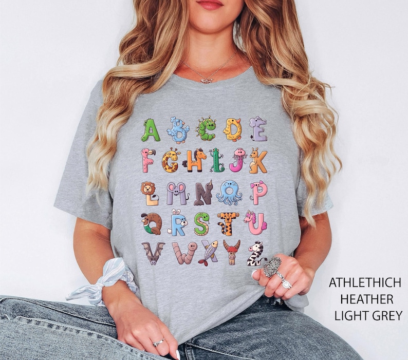K&ouml;nnte beinhalten: Hellgraues T-Shirt mit einem farbenfrohen Alphabet-Design, wobei jeder Buchstabe durch eine Tierillustration dargestellt wird. Das Shirt besteht aus einem weichen Material und eignet sich f&uuml;r Freizeitkleidung. Der Text "ATHLETHICH HEATHER LIGHT GREY" ist rechts aufgedruckt.