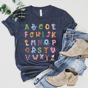K&ouml;nnte beinhalten: Dunkelblaues T-Shirt mit einem farbenfrohen Alphabet-Design, bei dem jeder Buchstabe als Tier dargestellt ist. Das Shirt wird mit blauen Jeans und beigen Stiefeletten kombiniert, was ein l&auml;ssiges, stilvolles Outfit ergibt.