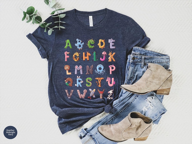 K&ouml;nnte beinhalten: Dunkelblaues T-Shirt mit einem farbenfrohen Alphabet-Design, bei dem jeder Buchstabe als Tier dargestellt ist. Das Shirt wird mit blauen Jeans und beigen Stiefeletten kombiniert, was ein l&auml;ssiges, stilvolles Outfit ergibt.