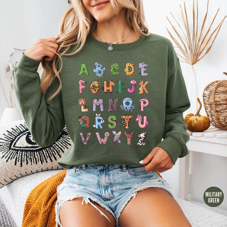 K&ouml;nnte beinhalten: Olivgr&uuml;nes Sweatshirt mit einem farbenfrohen Alphabet-Design. Jeder Buchstabe ist kreativ mit Tiermotiven und Details illustriert. Das Sweatshirt ist ein bequemer, l&auml;ssiger Stil.