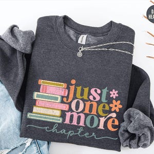 May include: Dark heather gray sweatshirt with the text "just one more chapter" in colorful letters and a stack of books. A silver necklace with a circular pendant is draped across the sweatshirt. The sweatshirt is folded on top of a pair of blue jeans.