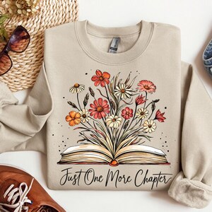 May include: A beige sweatshirt with a floral design and an open book with the text "Just One More Chapter".